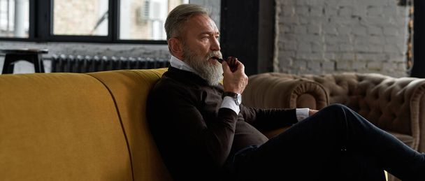 Senior thoughtful caucasian man smoking tobacco pipe on sofa in spacious apartment. Stylish bearded pensioner wearing casual clothes. Modern elderly male lifestyle. Home rest and free time - Foto, Imagem