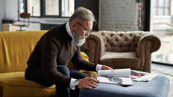 Senior focused caucasian man drinking whiskey and reading journal on sofa in spacious apartment. Stylish bearded pensioner wearing glasses. Modern elderly male lifestyle. Domestic rest and leisure - Zdjęcie, obraz