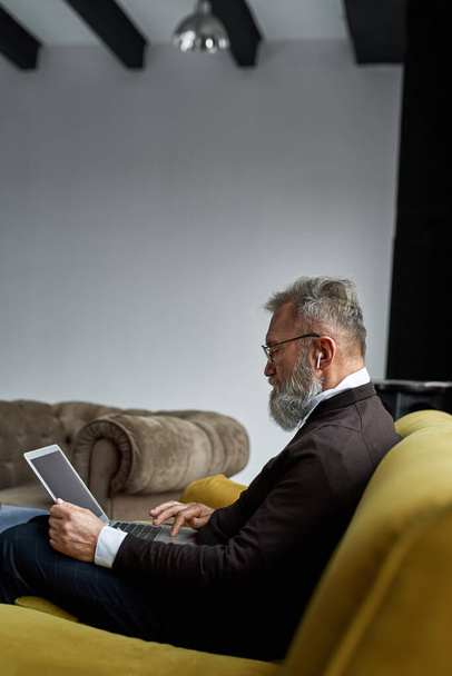 Elderly caucasian man using and watching laptop on sofa at home. Trendy bearded pensioner wearing glasses and wireless earphones. Modern senior male lifestyle. Domestic entertainment and leisure - Фото, изображение