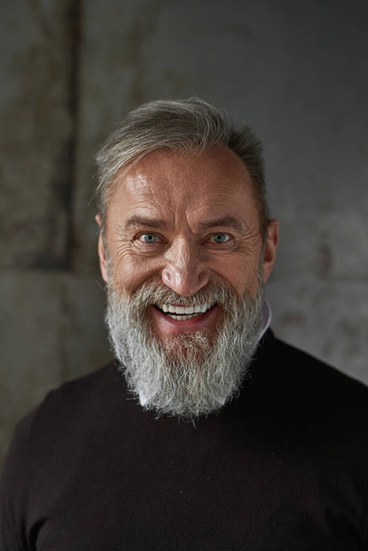 Portrait of grey hair smiling caucasian man looking at camera. Trendy pensioner wearing sweater. Concept of modern elderly male lifestyle. Isolated on rusty concrete background. Studio shoot - Фото, зображення