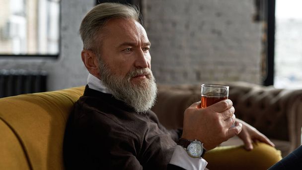 Senior thoughtful caucasian man drinking whiskey or brandy on sofa in spacious apartment. Stylish bearded pensioner wearing casual clothes. Modern elderly male lifestyle. Home rest and free time - Zdjęcie, obraz