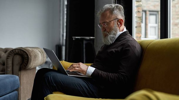 Senior caucasian man typing on laptop on sofa in spacious apartment. Stylish bearded pensioner wearing glasses and wireless earphones. Modern senior male lifestyle. Domestic entertainment and leisure - Foto, Bild