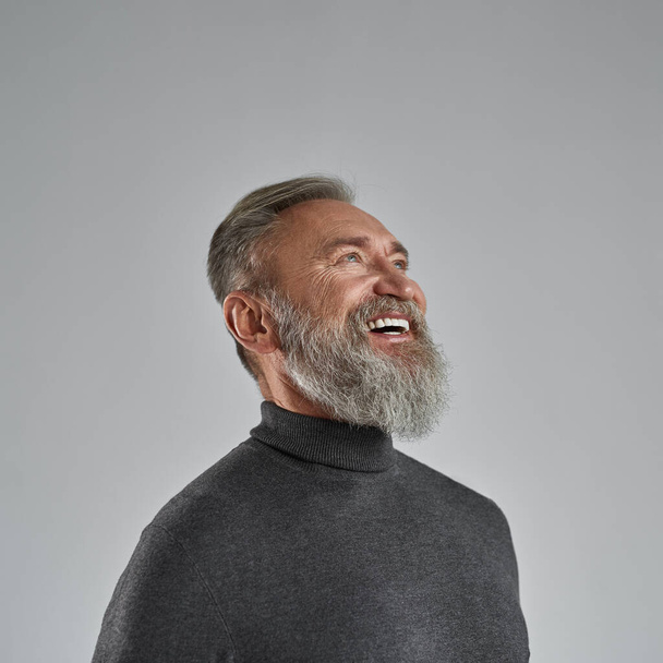 Cropped image of grey hair smiling caucasian man looking up. Fashionable bearded pensioner wearing sweater. Concept of modern elderly male lifestyle. Isolated on grey background in studio. Copy space - Foto, Imagem