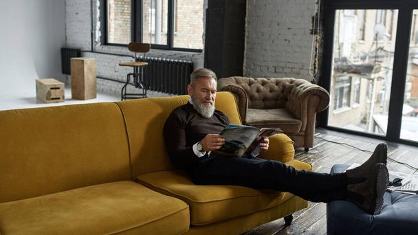 Grey hair concentrated caucasian man reading journal on sofa at home. Fashionable bearded pensioner. Modern senior male lifestyle. Domestic rest and leisure. Spacious apertment flat. Daytime - Foto, immagini
