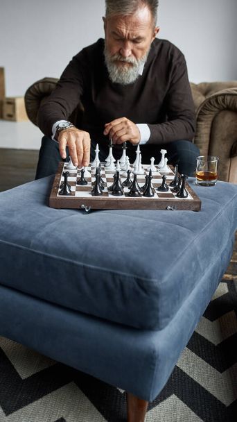 Elderly thoughtful caucasian man playing chess with himself in armchair at home. Elegant bearded pensioner. Modern elderly male lifestyle. Domestic entertainment, hobby and leisure. Spacious apartment - Photo, image