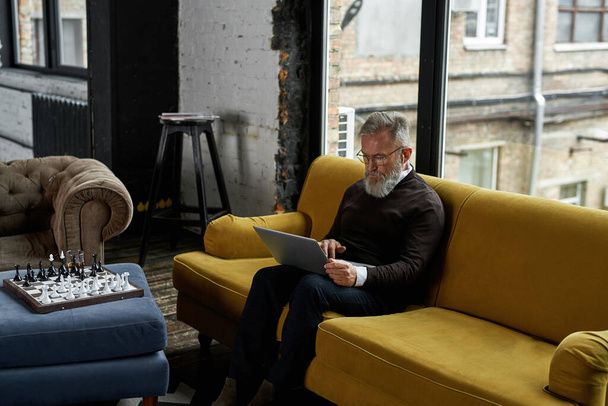 Grey hair caucasian man using and watching laptop on sofa in spacious flat. Bearded pensioner wearing glasses and wireless earphones. Modern senior male lifestyle. Domestic entertainment and leisure - Zdjęcie, obraz