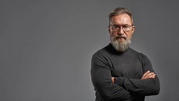 Confident senior caucasian man with crossed arms looking at camera. Stylish bearded pensioner wearing sweater and glasses. Modern elderly male lifestyle. Grey background. Studio shoot. Copy space - Φωτογραφία, εικόνα