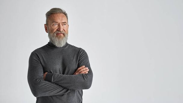 Senior serious caucasian man with crossed arms looking away. Stylish bearded pensioner wearing sweater. Concept of modern elderly male lifestyle. Isolated on white background. Studio shoot. Copy space - Fotó, kép