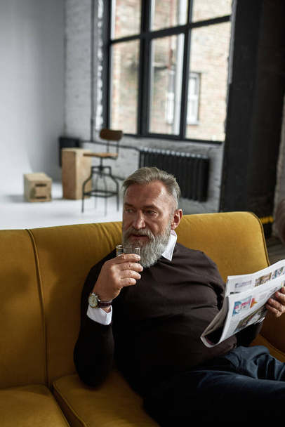 Grey hair thoughtful caucasian man drinking cognac and reading newspaper on sofa in spacious flat. Fashionable bearded pensioner. Modern senior male lifestyle. Domestic rest and leisure - Фото, зображення
