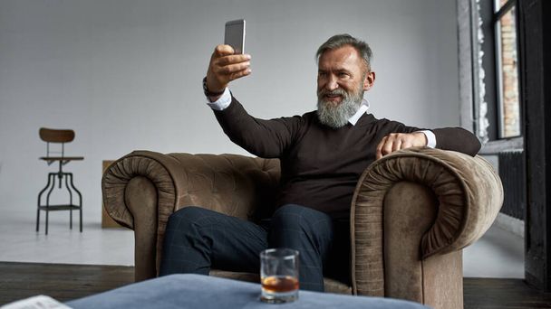 Elderly smiling elegant caucasian man taking selfie on smartphone in armchair in spacious apartment. Trendy bearded pensioner. Modern senior male lifestyle. Domestic entertainment and leisure - Φωτογραφία, εικόνα