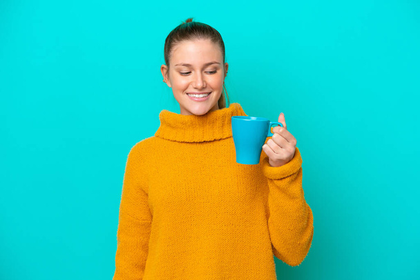 Молода біла жінка тримає чашку ізольовано на синьому фоні з щасливим виразом
 - Фото, зображення