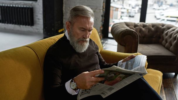 Grey hair concentrated caucasian man reading newspaper on sofa in spacious flat. Fashionable bearded pensioner wearing casual clothes. Modern senior male lifestyle. Domestic rest and leisure - Foto, Imagem