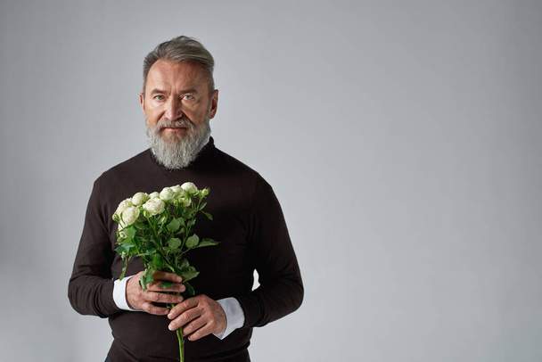 Elderly serious caucasian man with bouquet of white roses. Bearded male wearing sweater. Present for romantic meeting, Vaentines Day and 8 march. Isolated on grey background. Studio shoot. Copy space - Foto, imagen