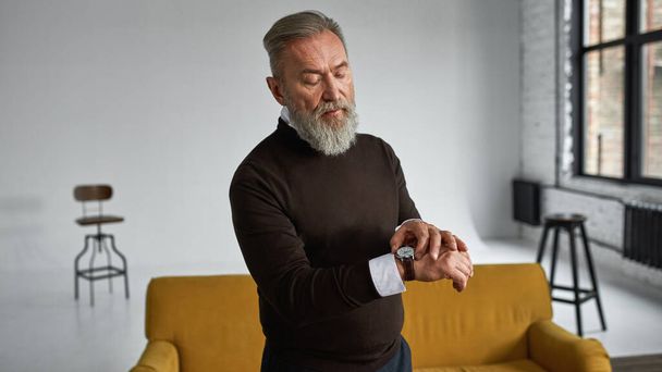 Grey hair focused caucasian man watching time on wristwatch in spacious apartment. Fashionable bearded pensioner wearing casual clothes. Concept of modern elderly male lifestyle. Daytime - Foto, Bild