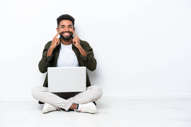 Young Brazilian man with a laptop sitting on the floor isolated on white smiling with a happy and pleasant expression - Fotografie, Obrázek