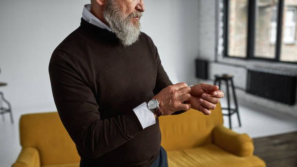 Obscure face of senior serious man correcting shirt sleeve in spacious apartment. Stylish bearded pensioner wearing casual clothes. Concept of modern elderly male lifestyle. Daytime - Photo, image