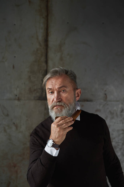 Senior caucasian man combing his beard with comb. Stylish pensioner. Modern elderly male lifestyle. Personal beauty and hygiene. Isolated on rusty concrete background. Studio shoot. Copy space - Фото, зображення