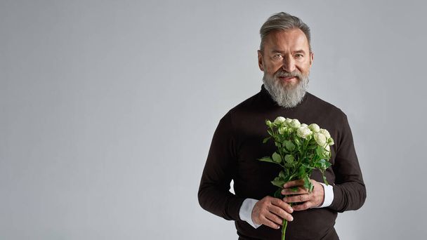 Senior european man with bouquet of white roses looking at camera. Stylish bearded male. Present for romantic meeting, Vaentines Day and 8 march. Isolated on grey background. Studio shoot. Copy space - Photo, Image