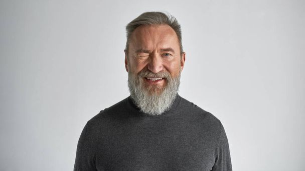 Elderly smiling caucasian man winking and looking at camera. Trendy bearded pensioner wearing sweater. Concept of modern senior male lifestyle. Isolated on white background. Studio shoot. Copy space - Φωτογραφία, εικόνα