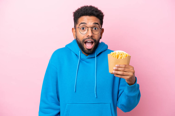 Young Brazilian man holding fried chips isolated on pink background with surprise and shocked facial expression - Zdjęcie, obraz