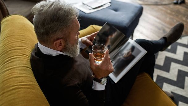 Top view of grey hair concentrated man drinking whiskey or brandy and reading journal on sofa in spacious flat. Fashionable bearded pensioner. Modern senior male lifestyle. Domestic rest and leisure - Fotó, kép