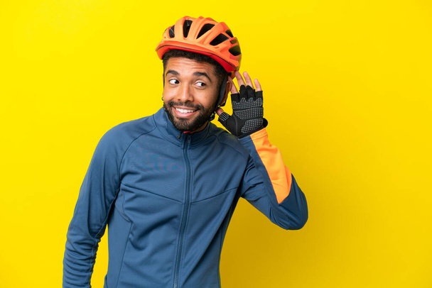 Young cyclist Brazilian man isolated on yellow background listening to something by putting hand on the ear - Valokuva, kuva
