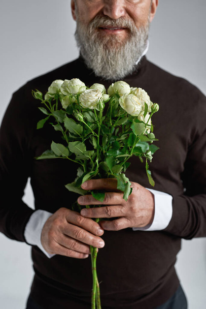 Bouquet of white roses in hands of cropped elderly man. Trendy bearded pensioner wearing sweater. Present for romantic meeting, Vaentines Day and 8 march. Isolated on grey background. Studio shoot - Фото, зображення