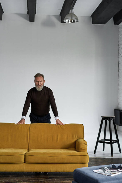 Elderly serious caucasian man standing by sofa and looking at camera in spacious apartment. Trendy bearded pensioner wearing casual clothes. Concept of modern senior male lifestyle. Daytime - Photo, image