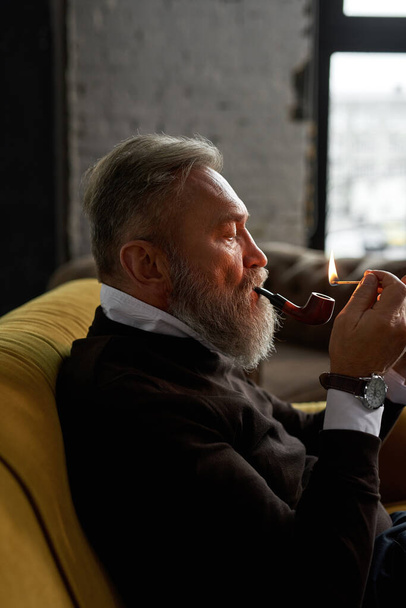 Senior focused caucasian man lighting tobacco pipe with match on sofa in spacious apartment. Stylish bearded pensioner wearing casual clothes. Modern elderly male lifestyle. Home rest and free time - Fotó, kép