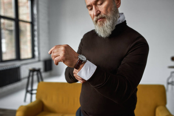 Cropped image of grey hair serious caucasian man correcting shirt sleeve in spacious flat. Fashionable bearded pensioner wearing casual clothes. Concept of modern elderly male lifestyle. Daytime - Фото, зображення
