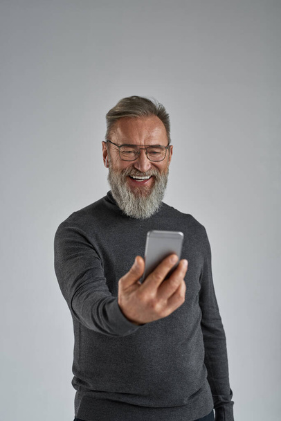 Senior smiling european man having video call on smartphone. Stylish pensioner wearing sweater, glasses and wireless earphones. Modern elderly male lifestyle. Grey background. Studio shoot. Copy space - 写真・画像