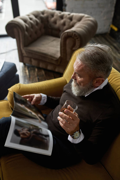 Top view of grey hair focused caucasian man drinking whiskey and reading journal on sofa in spacious flat. Fashionable bearded pensioner. Modern senior male lifestyle. Domestic rest and leisure - Foto, immagini
