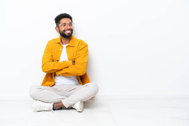 Young Brazilian man sitting on the floor isolated on white background with arms crossed and happy - 写真・画像