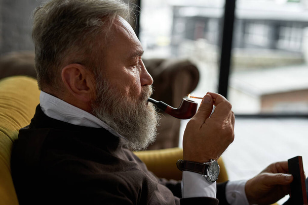 Grey hair focused caucasian man lighting tobacco pipe with match on sofa in spacious flat. Fashionable bearded pensioner wearing casual clothes. Modern senior male lifestyle. Home rest and free time - Фото, зображення