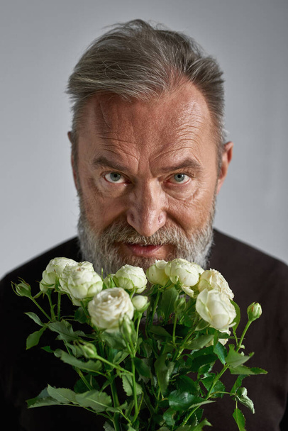 Portrait of grey hair serious european man with bouquet of white roses. Fashionable bearded male look at camera. Present for romantic meeting, Vaentines Day and 8 march. Grey background. Studio shoot - Foto, imagen