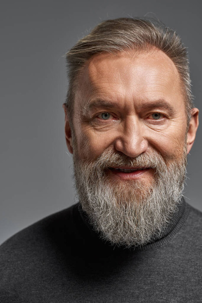 Portrait of smiling grey hair caucasian man looking at camera. Fashionable bearded pensioner wearing sweater. Concept of modern elderly male lifestyle. Isolated on grey background. Studio shoot - Photo, Image
