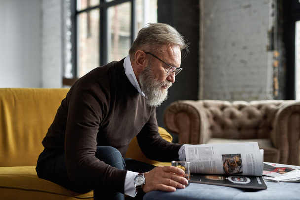 Elderly focused caucasian man drinking cognac and reading journal on sofa in spacious apartment. Trendy bearded pensioner wearing glasses. Modern senior male lifestyle. Domestic rest and leisure - Photo, Image