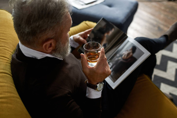 Top view of elderly concentrated man drinking cognac or brandy and reading journal on sofa in spacious apartment. Trendy bearded pensioner. Modern senior male lifestyle. Domestic rest and leisure - Foto, immagini