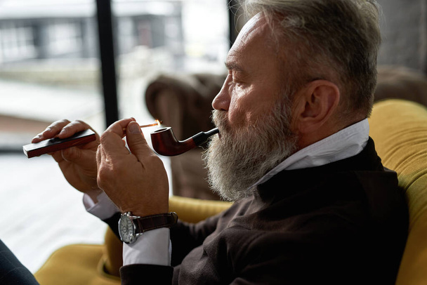 Elderly concentrated european man lighting tobacco pipe with match on sofa in spacious apartment. Trendy bearded pensioner wearing casual clothes. Modern senior male lifestyle. Home rest and free time - Foto, imagen