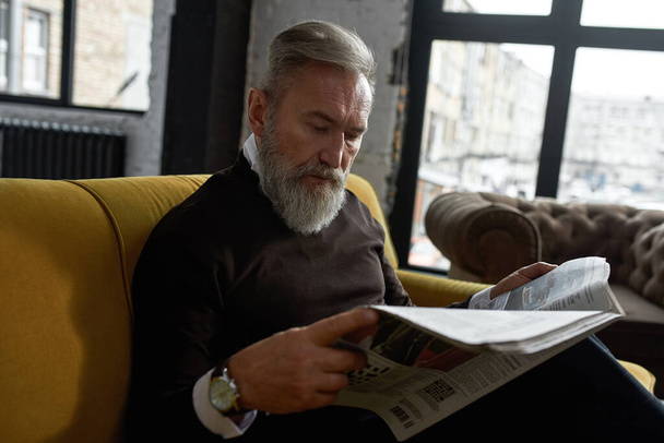 Senior focused caucasian man reading newspaper on sofa at home. Stylish bearded pensioner wearing casual clothes. Modern elderly male lifestyle. Domestic rest and leisure. Spacious apertment interior - Photo, Image