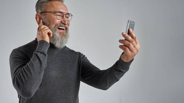 Elderly smiling european man pushing wireless earphones for choosing music on smartphone. Pensioner wearing sweater and glasses. Modern senior male lifestyle. Grey background. Studio shoot. Copy space - 写真・画像