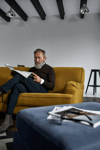 Senior concentrated caucasian man reading newspaper on sofa in spacious apartment. Stylish bearded pensioner wearing casual clothes. Modern elderly male lifestyle. Domestic rest and leisure - Fotografie, Obrázek