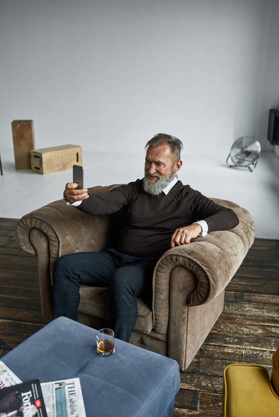 Grey hair smiling caucasian man taking selfie on smartphone in armchair in spacious flat. Fashionable bearded pensioner. Modern senior male lifestyle. Domestic entertainment and leisure - Фото, зображення