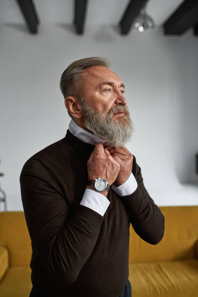 Partial image of ederly serious caucasian man correcting shirt collar in spacious apartment. Trendy bearded pensioner wearing casual clothes. Concept of modern senior male lifestyle. Daytime - Foto, Bild