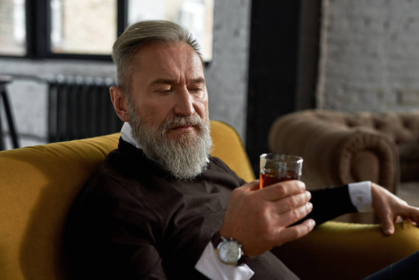 Grey hair serious caucasian man drinking and looking at glass with cognac or whiskey in his hand in spacious flat. Fashionable male sit on sofa. Modern senior male lifestyle. Home rest and free time - Φωτογραφία, εικόνα