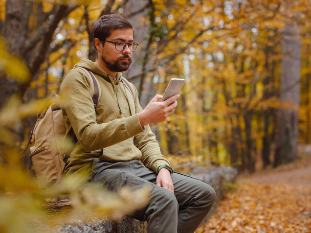 Νέος όμορφος άντρας στο φθινόπωρο δάσος με smartphone. νεαρός hipster τύπος με σακίδιο, ταξιδιώτης στέκεται στο δάσος, Πεζοπορία, Δάσος, Ταξίδι, ενεργό υγιεινό τρόπο ζωής, περιπέτεια, διακοπές έννοια. - Φωτογραφία, εικόνα