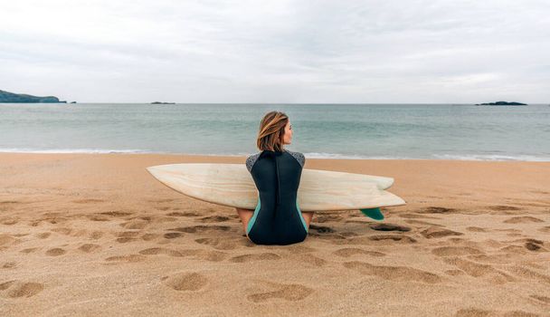 Jonge surfer vrouw met wetsuit en surfplank zittend op het zand opzij kijkend op het strand - Foto, afbeelding