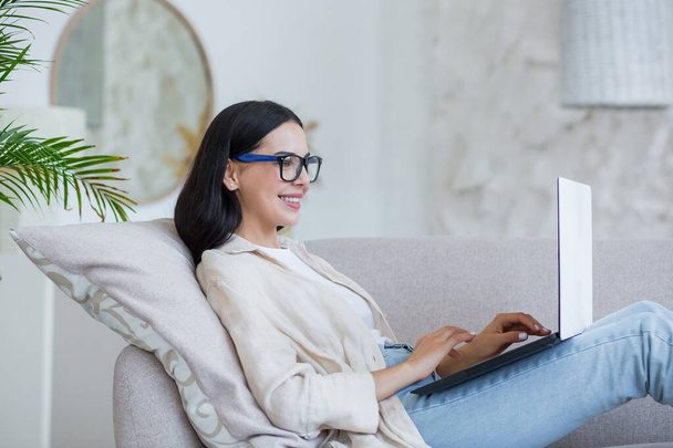Young beautiful girl studying online at home sitting on sofa in living room, brunette in glasses working with laptop remotely - Foto, Bild