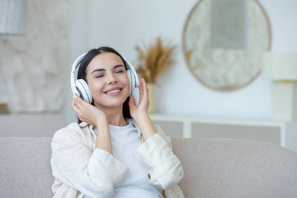 Beautiful young woman resting listening to music in white big headphones, brunette sitting on sofa in living room with closed eyes dreaming at home - Valokuva, kuva