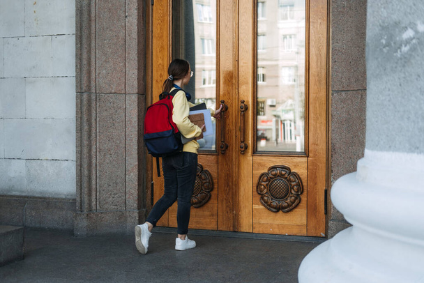 Студентська дівчина змішаної раси входить до дверей університету, коледжу. Портрет щасливої випадкової студентки з рюкзаком, ноутбуком та книгами на фоні університетських дверей
 - Фото, зображення